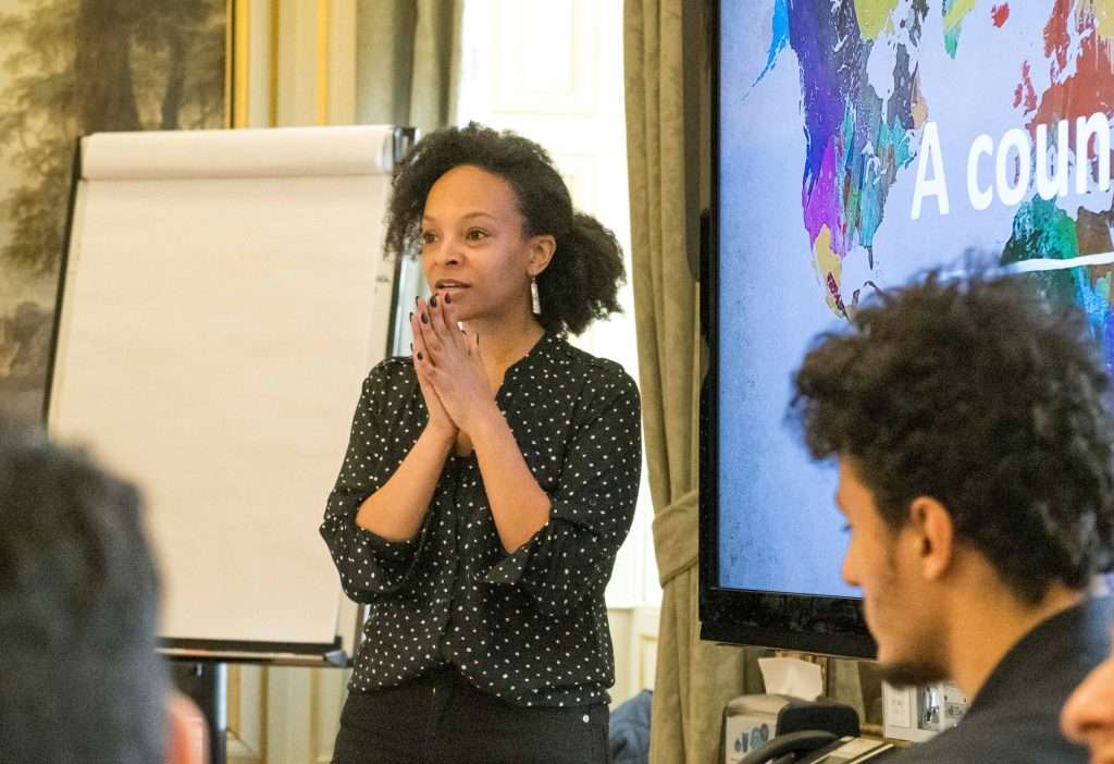 Black expat woman standing in front of audience with a white board and a screen behind her.